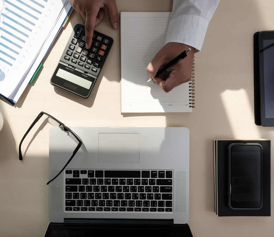 An overview image of a desk with a laptop, calculator, glasses, and paper.