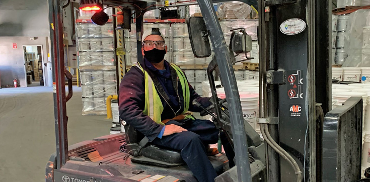 Man in construction vest and mask operating machinery in a warehouse.